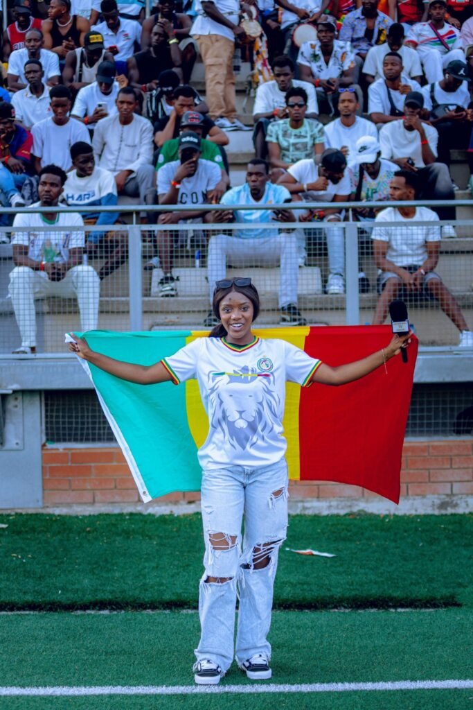 Young woman waves Cameroonian flag at vibrant outdoor sports event with cheering crowd.