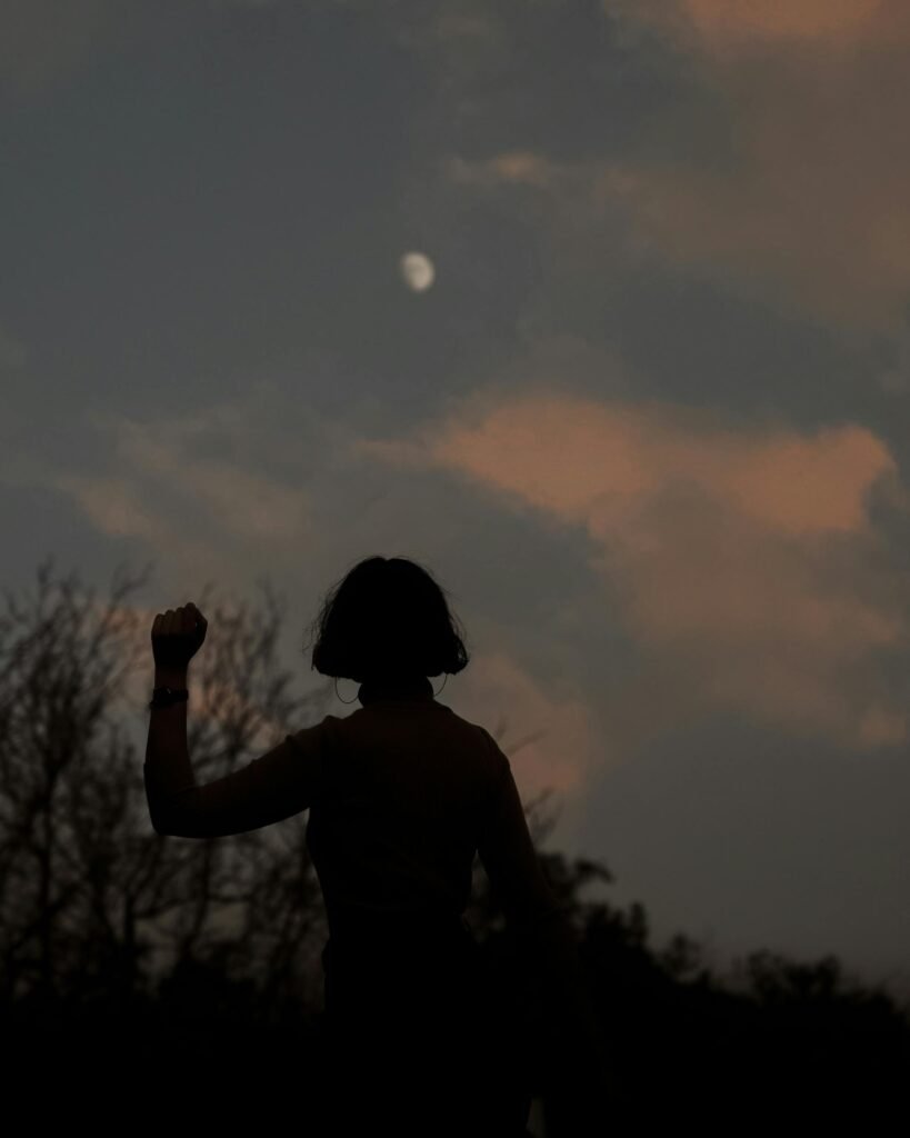pexels-photo-10032585-10032585 Silhouette of a person with a raised fist against a moonlit sky with scattered clouds.